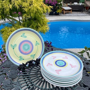 May include: A stack of white ceramic plates with a colorful design. Each plate has a central swirl of blue, pink, and yellow, surrounded by a ring of pink hearts or green leaves. The plates are stacked on a decorative metal table.