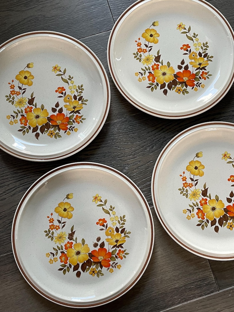 May include: Set of four white ceramic dinner plates with a brown rim. Each plate features a floral design with yellow, orange, and brown flowers.