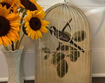 May include: A white birdcage with a bird illustration and a hook on top. The cage is decorated with a light green trim. The cage is sitting on a wooden surface with a vase of sunflowers in the background.