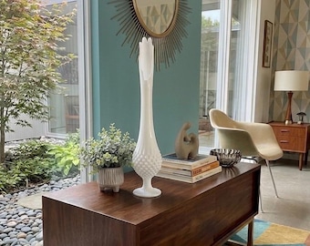 May include: A mid-century modern interior with a walnut cabinet on a gold metal frame. A white hobnail vase and a small potted plant sit on top. A starburst mirror hangs on a teal wall.
