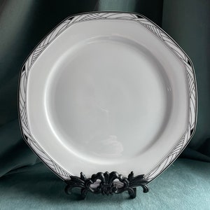 May include: White octagonal ceramic dinner plate with a black and white geometric design around the rim. The plate is on a black metal stand.