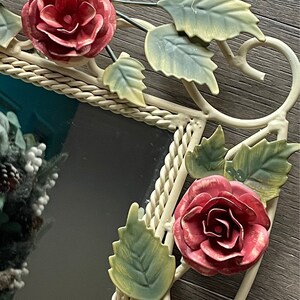 May include: A white metal mirror with a rope-like border and floral accents. The mirror is decorated with red roses and green leaves.