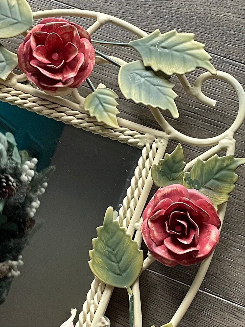 May include: A white metal mirror with a rope-like border and floral accents. The mirror is decorated with red roses and green leaves.
