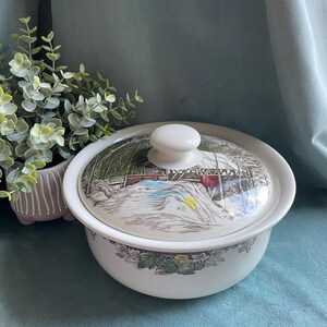 May include: A white ceramic serving bowl with a lid. The lid features a winter landscape scene with a bridge and snow. The bowl has a white knob and a decorative floral border. The bowl is approximately 8 inches in diameter.