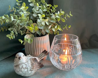 May include: A decorative arrangement featuring a clear glass candle holder with starburst designs, a lit candle, a ceramic bird figurine, and a potted plant with green foliage. The items are arranged on a teal surface.