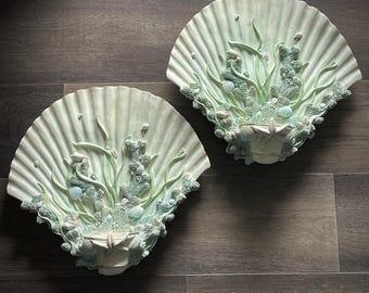May include: Two decorative wall plaques shaped like seashells with a 3D design of seaweed, seashells, and starfish. The seashells are a light green color with a white interior.