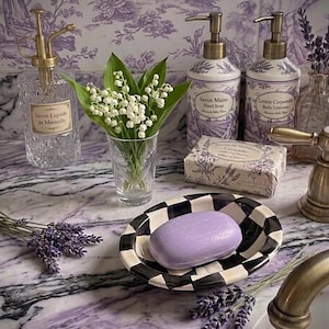 May include: A bathroom arrangement featuring lavender and white elements. It includes a black and white checkered soap dish with a purple soap bar, soap dispensers, a glass spray bottle, and a small vase with white flowers. Floral wallpaper in the background.