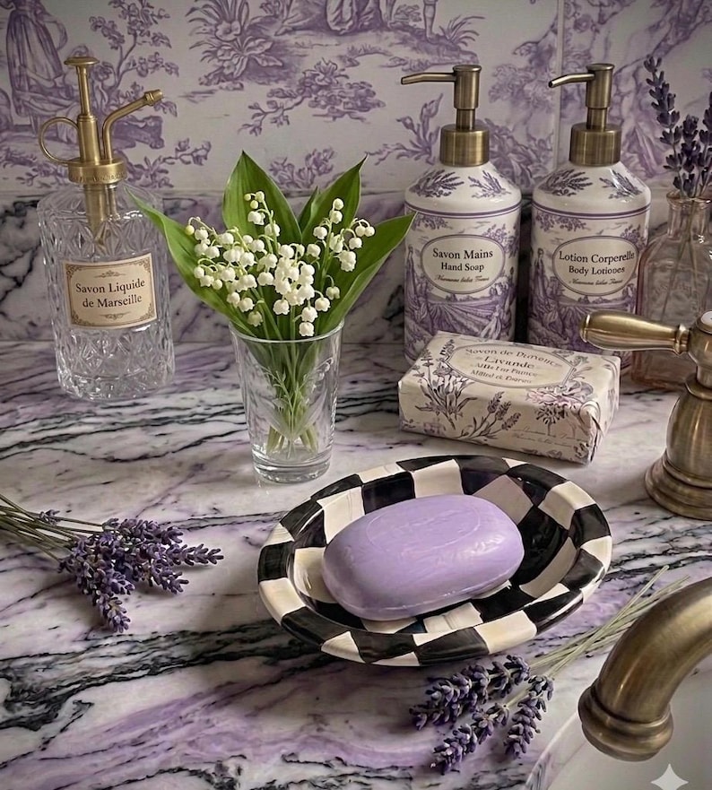 May include: A bathroom arrangement featuring lavender and white elements. It includes a black and white checkered soap dish with a purple soap bar, soap dispensers, a glass spray bottle, and a small vase with white flowers. Floral wallpaper in the background.