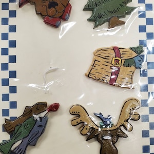 May include: A set of five handcrafted semi-stone buttons featuring a bear wearing a red hat, a green Christmas tree, a brown moose with a blue bird, a brown basket with two fish, and a brown fish. The buttons are packaged on a card with the text "Cache Junction Craftmart $4.97" and "Handcrafted Semi-Stone Buttons".