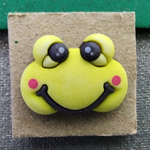 May include: A yellow frog face with black eyes and a black smile. The frog has pink cheeks.