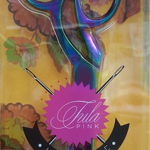 May include: A pair of iridescent rainbow-colored scissors with a black and white label that says "Tula Pink Hardware". The scissors are on a background of colorful floral fabric.