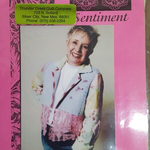 May include: A pink and black patterned fabric with a woman wearing a blue and pink patchwork jacket. The fabric has the text "Assembled Sentiment" and "vickitricks design".
