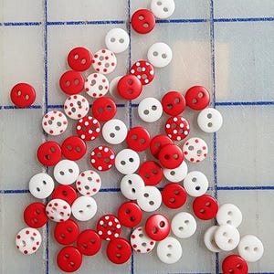 May include: A collection of small, round buttons in red and white. Some buttons are solid red, while others are white with red polka dots. The buttons have two holes for sewing and are scattered across a light blue grid.