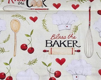 Op de afbeelding: Een witte stof met een crèmekleurige achtergrond met een herhalend patroon van rode harten, kersen, koksmutsen, deegrollers, garde en lepels. De tekst "Kiss the Cook" en "Bless the Baker" is in zwart gedrukt.