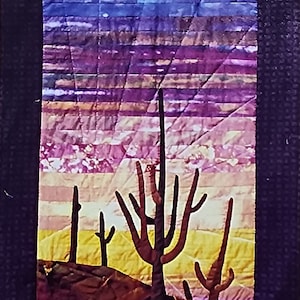 May include: A quilted wall hanging featuring a desert landscape with a sunset sky in shades of purple, pink, and orange. Silhouettes of tall cacti stand out against the colorful background.