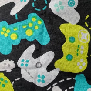 May include: Black fabric with a repeating pattern of colorful video game controllers. The controllers are in shades of green, blue, and white.