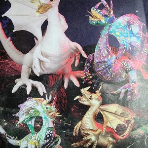 May include: Four plush dragon figures in various designs. One is solid white with gold accents, one features a vibrant multicolored pattern with purple and blue, one has a green and white floral print, and one is a metallic gold color with textured wings.