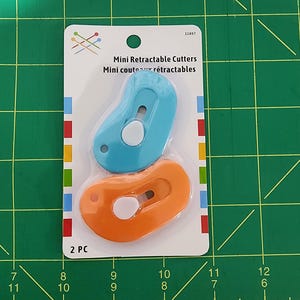 May include: A package containing two mini retractable cutters. One cutter is blue, and the other is orange. The package has the text "Mini Retractable Cutters" and "2 PC". The background is a green cutting mat.