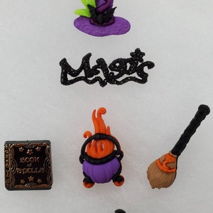 May include: A collection of Halloween-themed embellishments, including a witch's hat, the word "Magic", a cauldron, a broom, a book of spells, and a pair of high-heeled shoes. The items are in purple, orange, black, green, and gold.