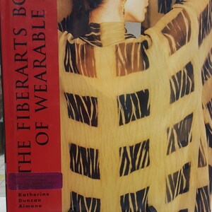 May include: A red book with the title "The Fiber Arts Book of Wearable Art" by Katherine Duncan Aimone. The cover features a woman wearing a brown and black zebra-print top.