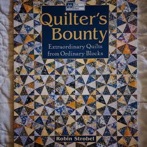 May include: A book titled "Quilter's Bounty: Extraordinary Quilts from Ordinary Blocks" by Robin Strobel. The cover features a colorful quilt pattern with blue, yellow, and white triangles.