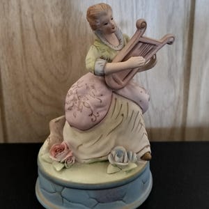 May include: A porcelain figurine of a woman playing a harp. She is wearing a pink and green dress with floral details. The figurine is on a base with a blue and green stone design and pink and blue roses.