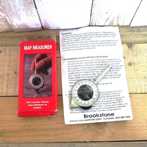 May include: A map measurer with a black and silver dial, packaged in a red box. The dial measures distance in inches. The box reads "MAP MEASURER" and "The counter shows map distance in inches."