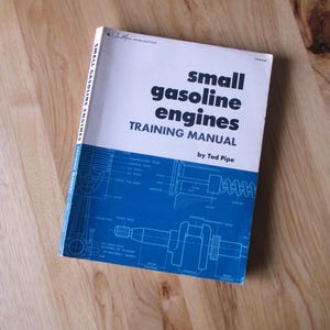 May include: A vintage training manual titled "Small Gasoline Engines" by Ted Pipe. The book has a white cover with blue accents and technical diagrams. The book is lying on a wooden surface.
