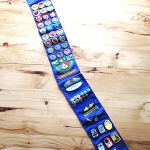 May include: A blue sash adorned with numerous colorful patches and badges, likely for a youth organization. The badges feature various designs and text, showcasing achievements and activities. The sash is displayed on a wooden surface.