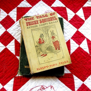 Puede incluir: Libro infantil vintage titulado "The Tale of Frisky Squirrel" de Arthur Scott Bailey, con ilustraciones de animales antropomórficos. El libro descansa sobre un libro verde y un edredón rojo y blanco.