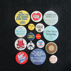 Könnte beinhalten: Eine Sammlung von runden Vintage-Anstecknadeln mit verschiedenen Logos und Slogans. Die Nadeln sind in verschiedenen Farben gehalten, darunter Rot, Blau, Gelb und Weiß. Einige der Logos sind "Love Pacer", "The Plymouth Voyager", "AMC Gremlin", "Cadillac Still #1", "We out sell "The other luxury car" 4 to 1", "The Cadillac Group", "Lease Me", "The Next Step Up", "I Can Be Very Friendly", "Bac America", "Riegle" und "Spirit".
