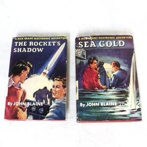 Pode incluir: Dois livros de capa dura vintage intitulados The Rocket's Shadow e Sea Gold, ambos parte da série Rick Brant Electronic Adventure de John Blaine. As capas apresentam ilustrações coloridas de pessoas, foguetes e cenas náuticas com texto em negrito.