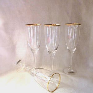 Puede incluir: Cuatro copas de champán transparentes con bordes dorados. Tres copas están de pie, mientras que una está tumbada de lado. Las copas tienen un diseño acanalado sobre un fondo blanco suave.