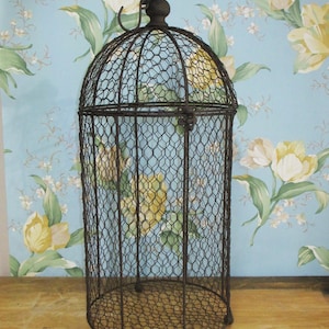 May include: A tall, cylindrical birdcage constructed from dark brown wire mesh with a hexagonal pattern. The cage features a domed top with a small decorative finial and a metal hanging loop. It stands on three small, rounded metal feet.