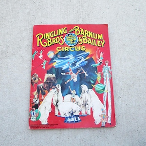 Peut inclure: Un magazine de programme vintage du cirque Ringling Bros. and Barnum & Bailey avec une couverture rouge illustrée sur le thème de l'espace, montrant une fusée, des ours polaires, un cheval, des acrobates et un artiste sur échasses sur un globe bleu.