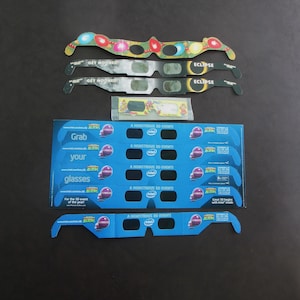 Könnte beinhalten: Eine Auswahl an 3D-Brillen und Verpackungen. Die Brillen haben farbenfrohe Rahmen mit Texten wie "GET MOONED!" und "ECLIPSE." Blaue Verpackungen zeigen "Grab your glasses" und "A MONSTROUS 3D EVENT!" mit dem Intel-Logo.