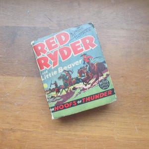 May include: A vintage paperback book titled "Red Ryder and Little Beaver on Hoofs of Thunder." The cover features a colourful illustration of cowboys riding horses. The book is part of the "Better Little Book" series.