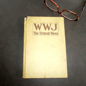 Könnte beinhalten: Ein Vintage-Buch mit dem Titel "WWJ The Detroit News" in braunen Buchstaben. Eine Brille mit Schildpattmuster liegt auf einer dunklen Oberfläche neben dem Buch. Das Buch hat ein abgenutztes, gealtertes Aussehen.