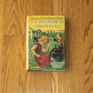 Pode incluir: Um livro de capa dura intitulado The Password to Larkspur Lane da série Nancy Drew Mystery Stories de Carolyn Keene. A sobrecapa amarela apresenta uma ilustração de uma pessoa com um vestido vermelho ajoelhada num jardim.