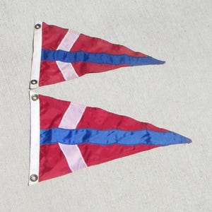 May include: Two triangular nautical pennant flags featuring a red background with a horizontal blue stripe and a vertical white stripe near the hoist. Each flag includes a white reinforced header strip along the left edge with two metal grommets.
