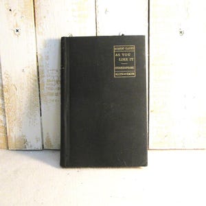 May include: A black book titled "As You Like It" by Shakespeare. The book has gold lettering and is part of the Academy Classics series, published by Allyn and Bacon. The book is set against a white and wood background.