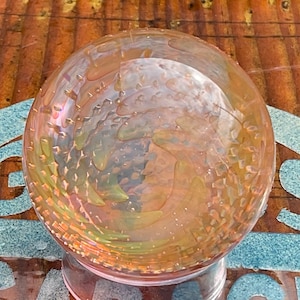 May include: A clear glass sphere with a swirling, iridescent pattern in shades of pink, orange, and yellow. The sphere is sitting on a wooden surface.