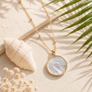 May include: A gold-colored necklace with a round pendant featuring a mother-of-pearl design of a sun and moon. The necklace is displayed on a sandy surface with a seashell and palm frond.