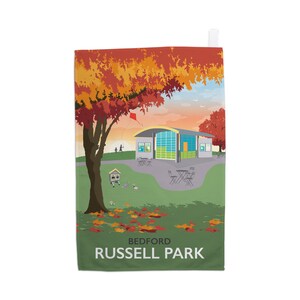以下が含まれることがあります： ベッドフォード・ラッセル公園の秋の風景を描いた装飾用タオル。曲がった屋根の建物、赤とオレンジの葉の木々、凧、公園の名前が白い文字でデザインされています。