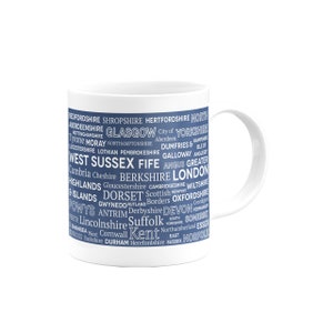 Puede incluir: Taza de cerámica blanca con fondo azul y texto blanco que enumera varios condados y regiones del Reino Unido. El texto está dispuesto en forma de nube de palabras.