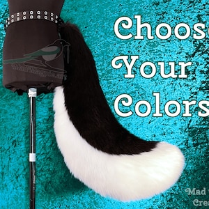 May include: A long, curved faux fur tail featuring a black upper section and a white lower section, attached to a black belt with metal eyelets. The text 'Choose Your Colors' is displayed in white lettering on the right side of the image.