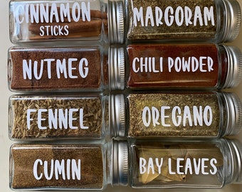 May include: Six glass spice jars with silver lids, each filled with different spices and labelled with white text. The labels read: Cinnamon Sticks, Marjoram, Nutmeg, Chilli Powder, Fennel, Oregano, Cumin, and Bay Leaves.