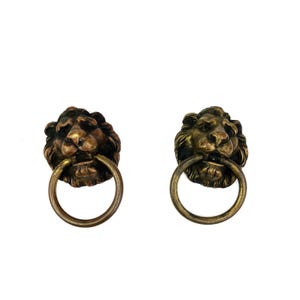May include: Two antique bronze lion head door knockers with ring handles. The lion heads have detailed manes and are mounted on a white background. The knockers are likely made of cast metal and are a decorative hardware item.