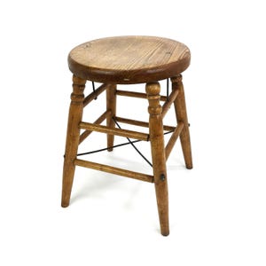 May include: A small, wooden stool with a round seat and four legs. The stool is made of light brown wood and has a classic design. The seat is approximately 12 inches in diameter.