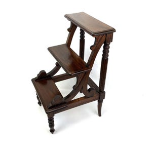 May include: A dark brown wooden library step stool featuring two tiers with a flat top surface. The structure is supported by four turned legs and decorative curved side panels that connect the steps, showcasing a polished wood grain finish.